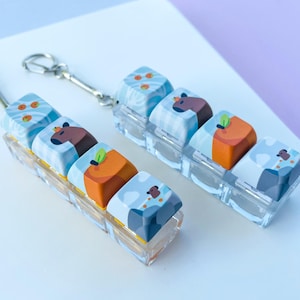 May include: Two keychains with custom keycaps. Each keycap features a unique design, including a brown bear, an orange fruit, and a blue and white pattern. The keycaps are mounted on a clear plastic base and attached to a silver keychain.
