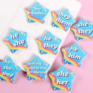May include: A set of nine star-shaped pronoun buttons with rainbow accents. The buttons feature various pronoun combinations, including "he/she", "they/them", "she/her", "he/him", and "any pronouns".
