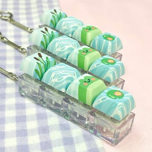 May include: A set of custom keycaps with a whimsical frog and water lily theme. The keycaps feature a variety of designs, including cattails, water patterns, and smiling frog faces. The keycaps are mounted on a clear plastic base.