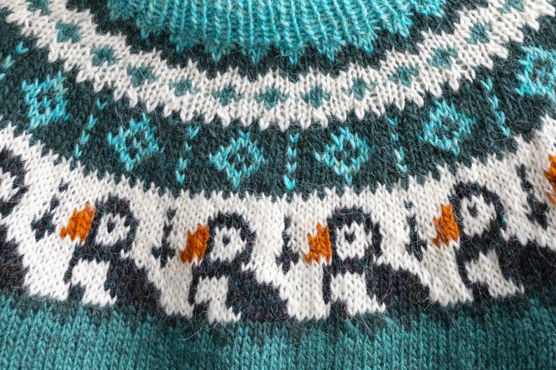A knitted jumper featuring a yoke with a repeating pattern of puffins in black, white, and orange, set against a white background. Above the puffins is a dark teal band with a white diamond motif and a ribbed teal neckline.