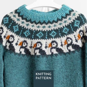 Könnte beinhalten: Ein blaugrüner Strickpullover mit Rundhalsausschnitt und einer Passe, die mit einem weißen Band verziert ist, das ein wiederkehrendes Muster aus schwarz-weißen Papageientauchern mit orangefarbenen Schnäbeln zeigt, gekrönt von einem geometrischen Design.