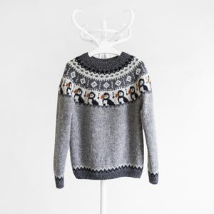 A grey knit jumper featuring a round neckline and a yoke decorated with a repeating pattern of black, white, and orange puffin birds, complemented by geometric diamond shapes in white and black against a dark grey background.