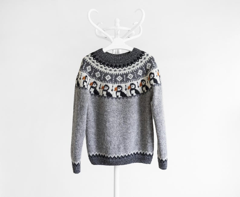 A grey knit jumper featuring a round neckline and a yoke decorated with a repeating pattern of black, white, and orange puffin birds, complemented by geometric diamond shapes in white and black against a dark grey background.