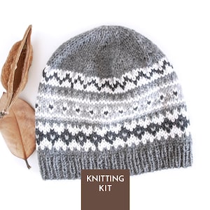 Puede incluir: Un gorro de punto gris, blanco y carbón con un patrón geométrico repetitivo y un borde acanalado. Una etiqueta rectangular marrón en la parte inferior muestra el texto blanco KNITTING KIT. El gorro está colocado junto a dos vainas de semillas secas.