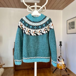 A teal knitted jumper featuring a round neckline and long sleeves. The yoke displays a detailed fair isle pattern with white, dark teal, and orange accents, including a repeating motif of small birds across the chest area.