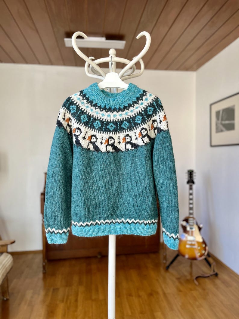 A teal knitted jumper featuring a round neckline and long sleeves. The yoke displays a detailed fair isle pattern with white, dark teal, and orange accents, including a repeating motif of small birds across the chest area.