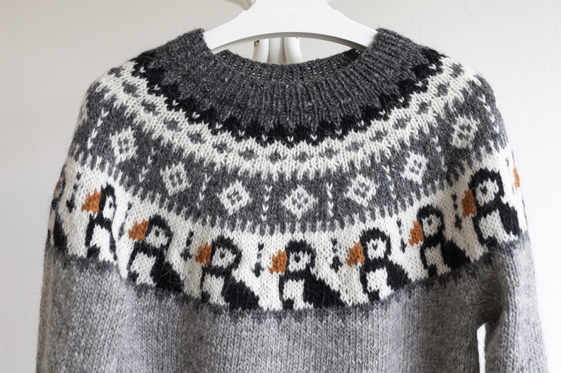 A grey knit jumper featuring a round neckline and a yoke decorated with a repeating pattern of black, white, and orange puffin birds, complemented by geometric diamond shapes in white and black against a dark grey background.