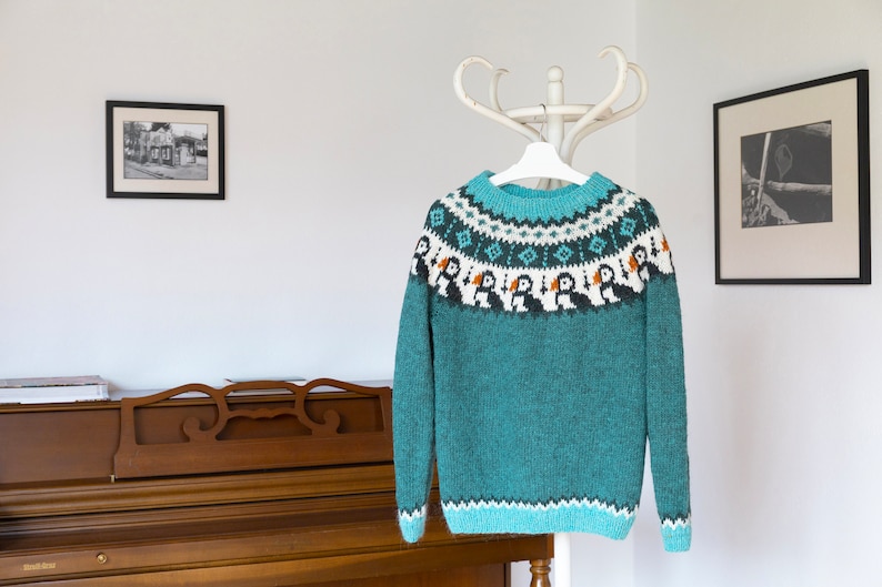 A teal long-sleeve knit jumper featuring a round neckline and a yoke decorated with a repeating pattern of black and white puffins with orange beaks, accented by geometric shapes in white and light blue, with a matching patterned hem.