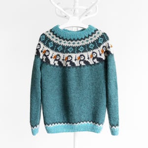 A teal knitted jumper featuring a circular yoke with a fair isle pattern. The design includes rows of geometric shapes in white and dark teal, and a row of stylised puffin birds with white bodies, dark wings, and orange beaks.