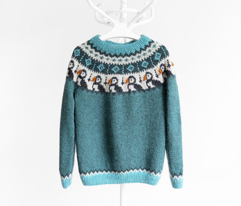 A teal knitted jumper featuring a circular yoke with a fair isle pattern. The design includes rows of geometric shapes in white and dark teal, and a row of stylised puffin birds with white bodies, dark wings, and orange beaks.