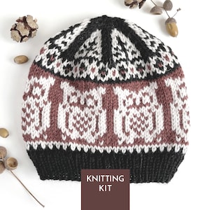 May include: A knitted beanie featuring a repeating white owl pattern on a dusty rose background, with a black ribbed brim and a black and white geometric design at the crown. A brown rectangular label at the bottom displays the text KNITTING KIT.