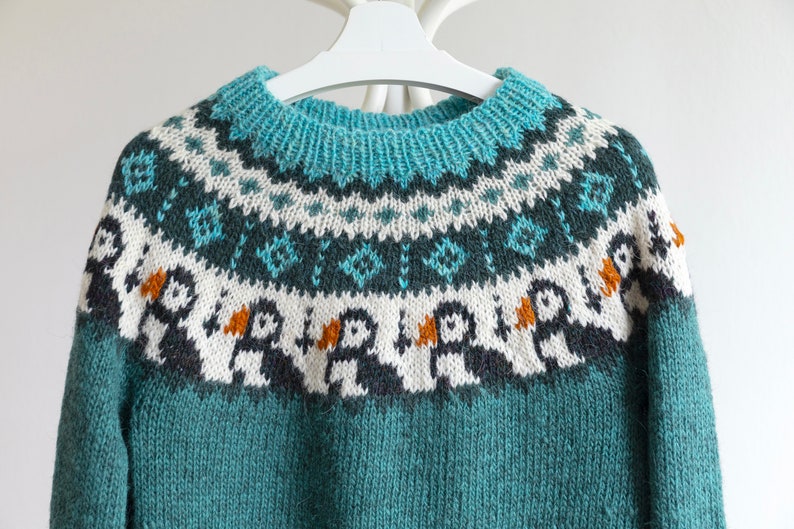 A teal knitted jumper featuring a circular yoke with a fair isle pattern. The design includes rows of geometric shapes in white and dark teal, and a row of stylised puffin birds with white bodies, dark wings, and orange beaks.