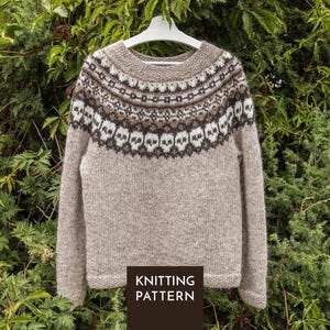 May include: A beige long-sleeve jumper featuring a round neckline and a yoke decorated with a repeating pattern of white skulls, dark brown, and black geometric shapes. A dark brown square at the bottom displays the text KNITTING PATTERN.