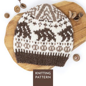 Può includere: Un berretto lavorato a maglia con un motivo fair isle con sagome di orsi marroni e bianchi, forme geometriche e un bordo a coste marrone scuro. Il berretto è mostrato con ghiande sparse e un'etichetta quadrata marrone con la scritta KNITTING PATTERN.