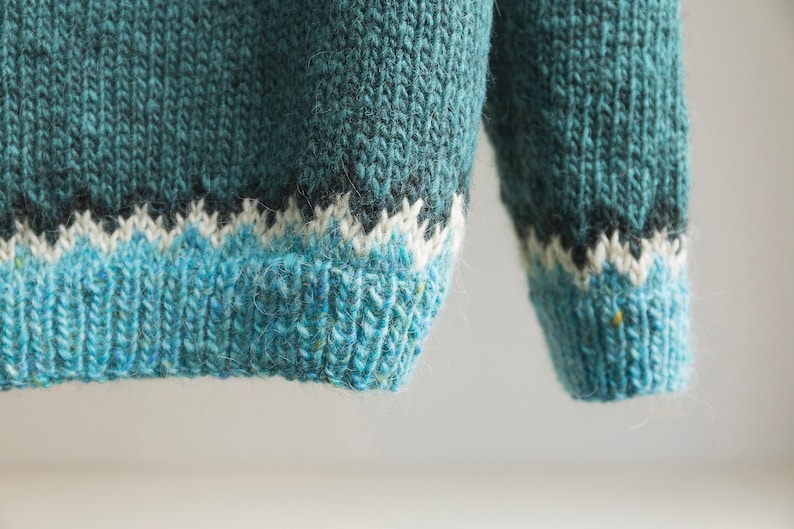 A long-sleeved knit jumper featuring a teal upper section, a dark charcoal band, a white zigzag pattern, and a light blue ribbed hem and cuff. The yarn has a textured appearance with subtle flecks of colour throughout the knit fabric.