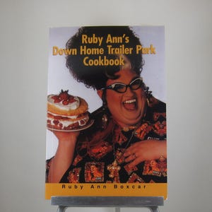 May include: A softcover cookbook titled Ruby Ann's Down Home Trailer Park Cookbook by Ruby Ann Boxcar. The cover features a person with a large dark hairstyle, cat-eye glasses, and a black shirt with Elvis prints, holding a strawberry shortcake.