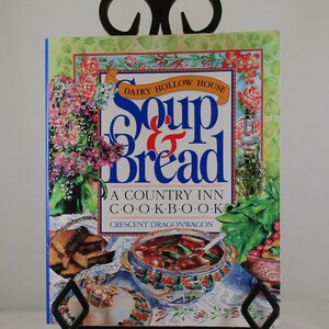 May include: A cookbook titled "Soup & Bread" is displayed on a black metal stand. The cover features a colorful illustration of soup, bread, and floral arrangements. The text on the cover reads "Dairy Hollow House" and "A Country Inn Cookbook."