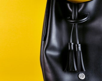May include: A black leather bucket bag with a drawstring closure and two tassel accents. The bag has a silver logo on the front.