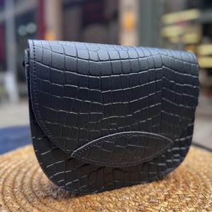 May include: Black crossbody bag with a crocodile-skin texture. The bag has a curved flap with a leaf-shaped accent. The bag is sitting on a woven, circular surface.