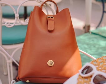 Può includere: Uno zaino in pelle marrone con chiusura a fibbia e un logo dorato. Lo zaino è seduto su una sedia blu.