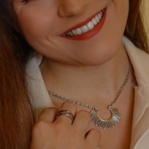 Puede incluir: Una mujer con cabello largo y castaño lleva una camisa blanca con un collar de plata con un diseño de sol. Lleva un anillo de plata en su dedo.