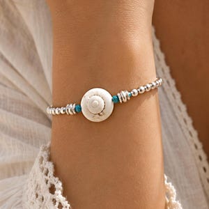 Op de afbeelding: Een kralenarmband met een centraal wit, cirkelvormig schelpbedeltje, geflankeerd door kleine turquoise kralen en metalen zilverkleurige tussenkralen. De armband is opgebouwd uit een reeks gepolijste zilverkleurige bolvormige kralen.
