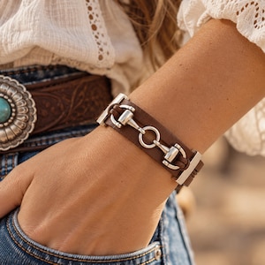 May include: A brown leather cuff bracelet featuring a silver-toned metal horse bit hardware centrepiece. The bracelet has a smooth, wide band design with metallic end caps that secure the leather strap, creating a structured look.