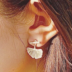 Puede incluir: Pendientes de plata con forma de hoja de ginkgo con poste de barra. Los pendientes se muestran en la oreja de una persona.