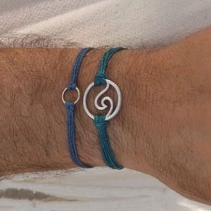 Puede incluir: Dos pulseras en una muñeca. Una pulsera es azul turquesa con un dije de ola plateado. La otra pulsera es azul oscuro con un dije de círculo plateado.