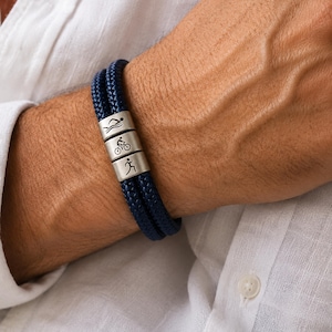 May include: A navy blue braided bracelet with three silver beads. Each bead features a small engraved symbol representing swimming, cycling, and running. The bracelet is worn on a wrist, showcasing a casual style.