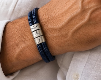 May include: A navy blue braided bracelet with three silver beads. Each bead features a small engraved symbol representing swimming, cycling, and running. The bracelet is worn on a wrist, showcasing a casual style.