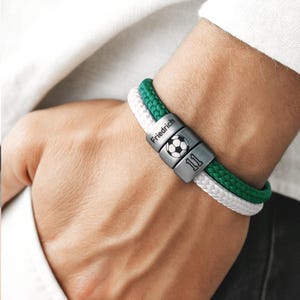 Könnte beinhalten: Ein personalisiertes Armband mit einem Fußball-Design und der Nummer 11. Das Armband hat ein grün-weißes Seil-Design mit einem silbernen Verschluss, auf dem der Name "Friedrich" eingraviert ist.