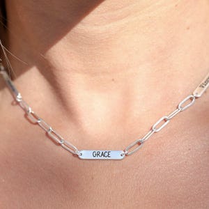 Op de afbeelding: Een zilveren ketting met een rechthoekige hanger met de tekst "GRACE" in zwarte letters. De ketting heeft een schakelkettingontwerp en wordt om de nek gedragen.