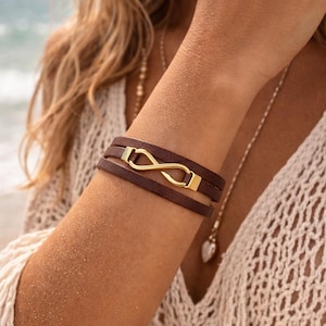 May include: A brown leather wrap bracelet with a gold-colored infinity symbol clasp. The bracelet has three leather straps and a rectangular gold clasp. The jewelry is worn on the wrist.