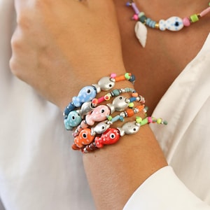 Könnte beinhalten: Eine Ansammlung farbenfroher Armbänder mit silbernen Fischanhängern. Die Armbänder sind in Blau-, Rosa-, Orange- und Rottönen gehalten und mit grünen und orangen Perlen akzentuiert. Eine passende Halskette mit einem weißen Muschelanhänger ist ebenfalls zu sehen.