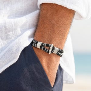 Puede incluir: Una pulsera de cuerda en blanco y negro con cierres de metal plateado. La pulsera se lleva en la muñeca, con una camisa blanca y pantalones azul marino en el fondo. La pulsera es de estilo náutico.