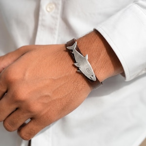 Op de afbeelding: Een bruine leren armband met een zilverkleurige metalen haaienbedel. De haaienbedel is horizontaal in het midden van de leren band geplaatst en toont een gedetailleerd silhouet met zichtbare kieuwspleten en een puntige rugvin.