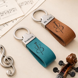 Può includere: Due portachiavi in pelle con anelli e finiture in metallo color argento. Uno è marrone chiaro con il nome Ms. Katie e l'illustrazione di un violoncello, l'altro è verde acqua con il nome Frederick e l'illustrazione di un violoncello.