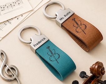 Puede incluir: Dos llaveros de cuero con anillas y herrajes de metal plateado. Uno es de color tostado con el nombre Ms. Katie y una ilustración de un violonchelo, el otro es de color verde azulado con el nombre Frederick y un violonchelo grabado.