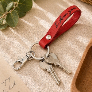 May include: Red leather keychain with the phrase "I love you" in black script. The keychain has a silver clasp and ring, holding three keys. A silver snap secures the leather strap.