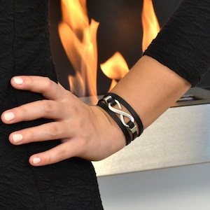 Könnte beinhalten: Schwarzes Lederarmband mit einem silbernen Unendlichkeitszeichen. Das Armband hat mehrere Stränge und einen Verschluss. Das Unendlichkeitszeichen ist der Blickfang und verleiht Eleganz.