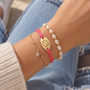 Op de afbeelding: Een set van drie armbanden bestaande uit een witte parelketting met gouden kralen, een felroze koordarmband met een gouden ovale bedel met een uitgesneden Kreeft sterrenbeeld, en een fijne gouden kettingarmband met kleine ronde gouden bedels.