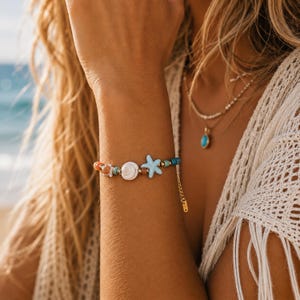 Könnte beinhalten: Ein Armband mit Perlen, das eine Muschel, einen Seestern und verschiedene bunte Perlen zeigt. Das Armband hat eine goldfarbene Kette und einen Verschluss. Der Schmuck wird am Handgelenk getragen, mit einem Strand- und Ozeanhintergrund.