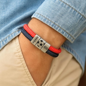 Può includere: Un braccialetto in pelle intrecciata rossa e blu navy con chiusura argentata. La chiusura presenta il nome "PIERRE", una grafica di pallone da calcio e il numero "9". Il braccialetto è indossato al polso.