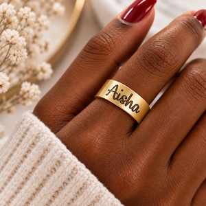 Peut inclure: Une bague anneau de couleur dorée avec le prénom Aisha gravé en écriture cursive noire. La bague présente une finition mate lisse et un design à surface large et plate qui entoure le doigt, mettant en valeur une inscription claire et lisible.