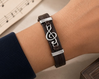 May include: A dark brown leather bracelet featuring a silver-toned metal treble clef charm centered between two silver-toned metal rectangular bars. The bracelet consists of multiple thin leather strands held together by the metal components.