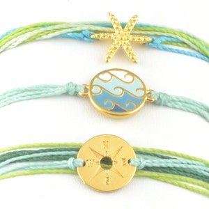 Puede incluir: Tres pulseras coloridas con dijes de oro. La pulsera superior tiene un dije de estrella de mar de oro, la pulsera del medio tiene un dije de círculo de oro con olas azules y blancas, y la pulsera inferior tiene un dije de brújula de oro.