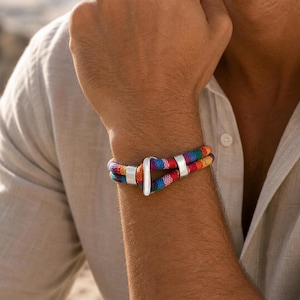 Op de afbeelding: Een kleurrijke touw armband met een zilveren sluiting. De armband heeft een levendige mix van rode, blauwe, gele en paarse draden. De zilveren sluiting heeft een eenvoudig ontwerp.