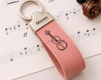 Puede incluir: Un llavero de cuero rosa con una pieza superior de metal plateado grabada con el nombre Maren. La correa de cuero presenta un dibujo lineal negro de un violín y un arco. El llavero está unido a una anilla metálica circular plateada.