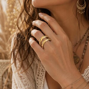 Op de afbeelding: Een goudkleurige ring met een dubbelbandontwerp. De ring wordt aan een hand gedragen, met andere gouden sieraden op de achtergrond. De gladde, gebogen banden van de ring creëren een moderne en elegante uitstraling. De sieraden maken waarschijnlijk deel uit van een mode- of accessoirecollectie.
